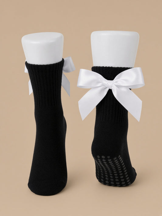 A pair of black ribbed ankle socks displayed on white mannequin legs against a solid beige background. One sock is shown from the front with a white satin bow tied around the ankle, and the other is shown from the back with a black grip sole.