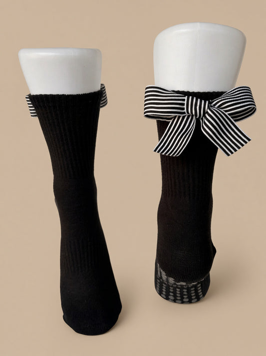 A pair of black ribbed crew socks displayed on white mannequin legs against a solid beige background. One sock is shown flat from the front, and the other is shown from the side with a black and white striped bow tied around the top.