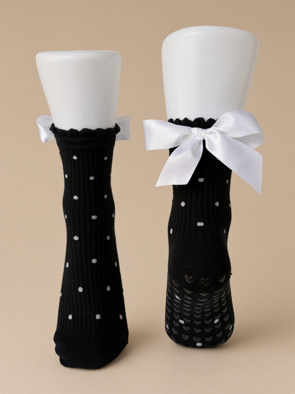A pair of black ribbed socks displayed on white mannequin legs against a warm beige background. One sock has small white polka dots, and the other features a large white satin bow at the top with a textured grip sole.