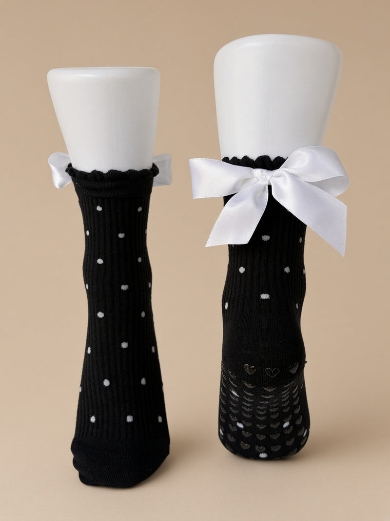 A pair of black ribbed socks displayed on white mannequin legs against a warm beige background. One sock has small white polka dots, and the other features a large white satin bow at the top with a textured grip sole.