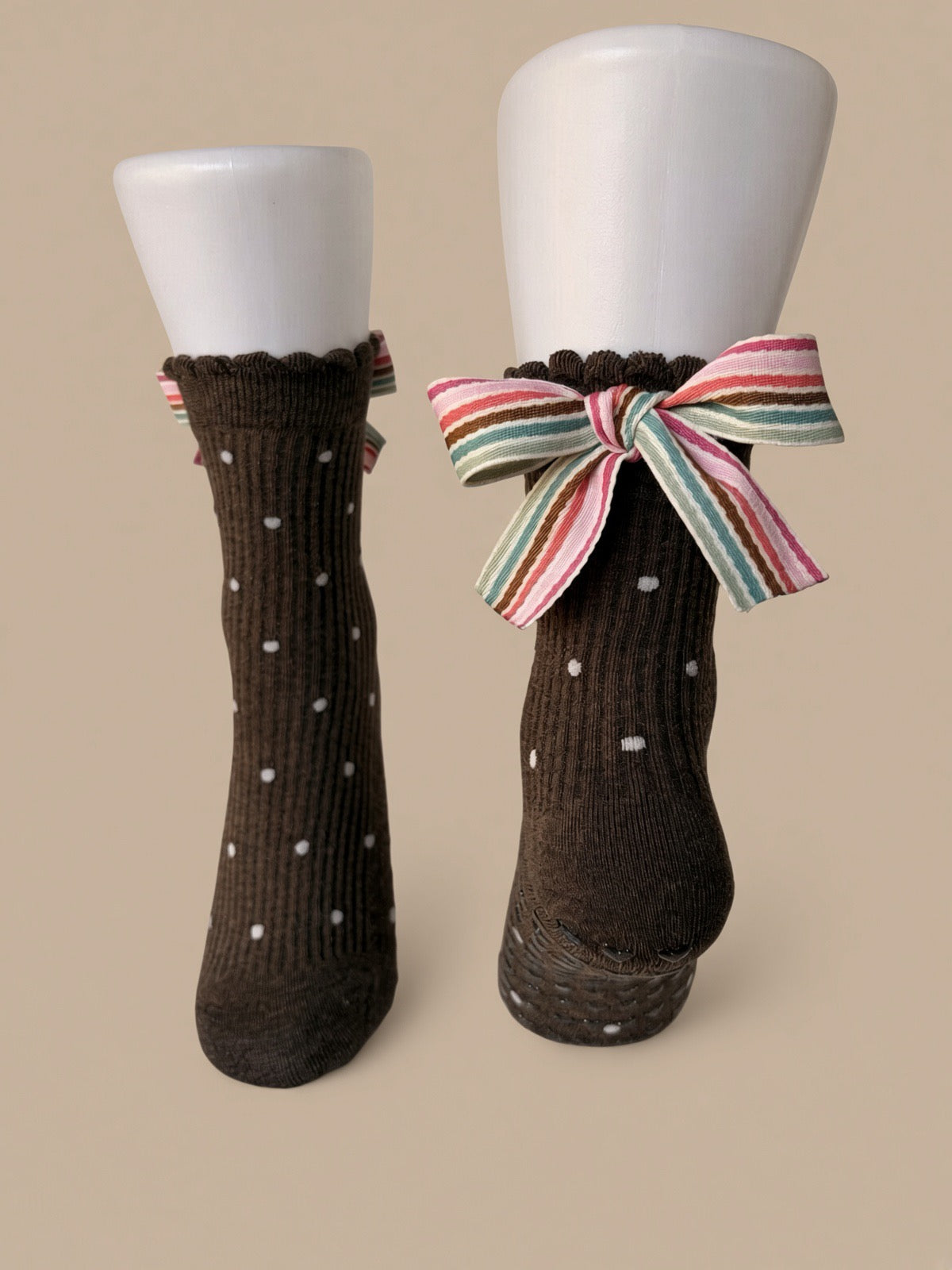 A pair of dark brown ribbed socks with small white polka dots, displayed on white mannequin legs. One sock is shown flat from the front, and the other is shown from the side/back, featuring a large striped bow in pink, green, and white tied around the ankle.