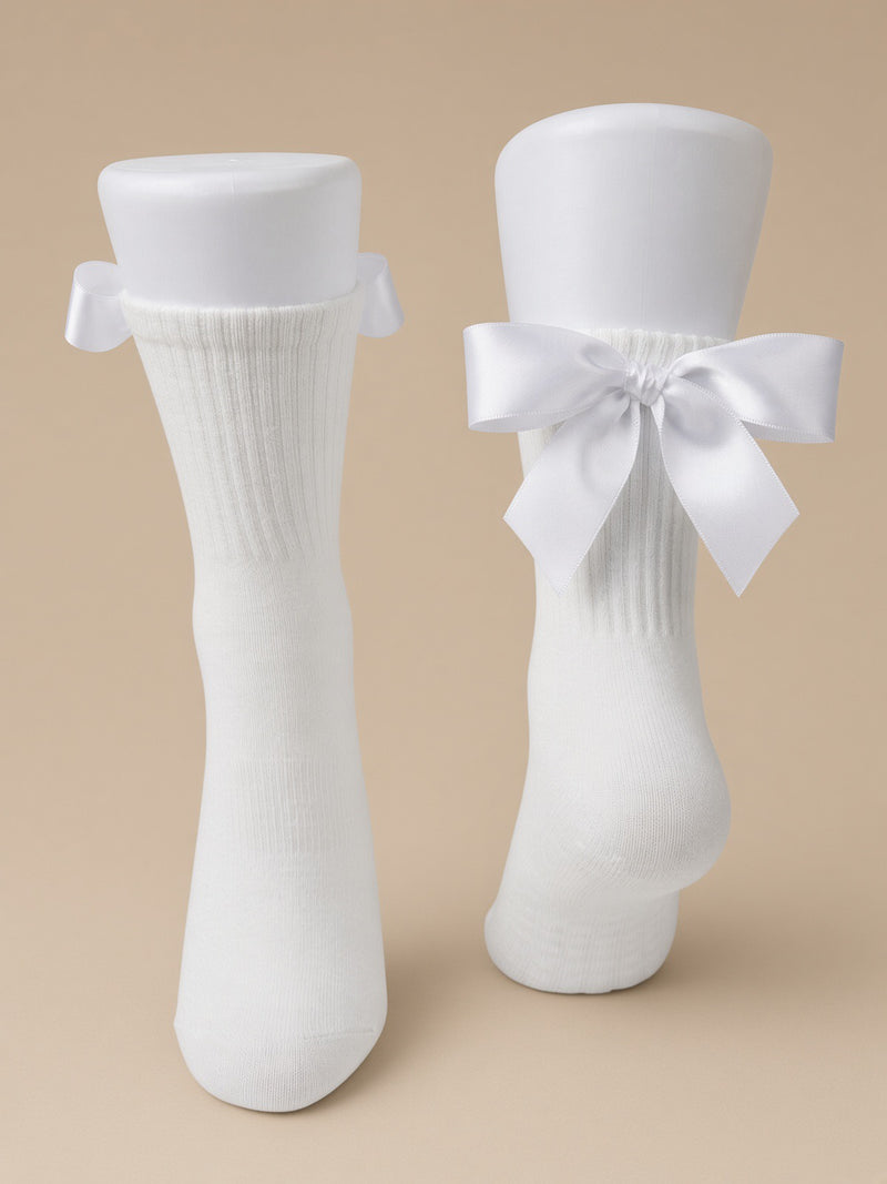 A pair of white ribbed crew socks displayed on mannequin legs against a warm beige background. One sock has a small bow at the top, and the other has a large bow at the ankle.