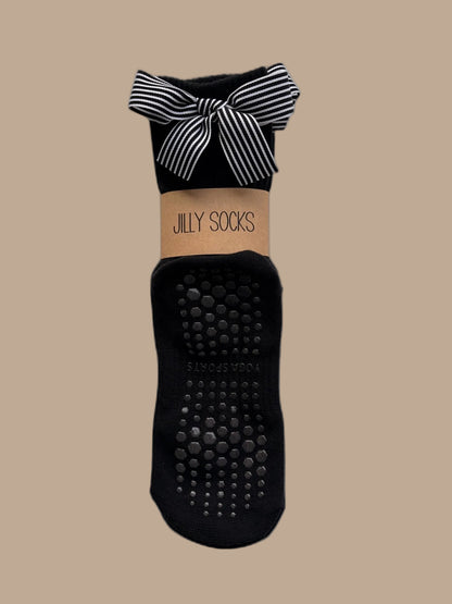 Cute, fashionable black Pilates grip socks with a black and white stripes bow and a tan paper band reading 'JILLY SOCKS' on a white background.